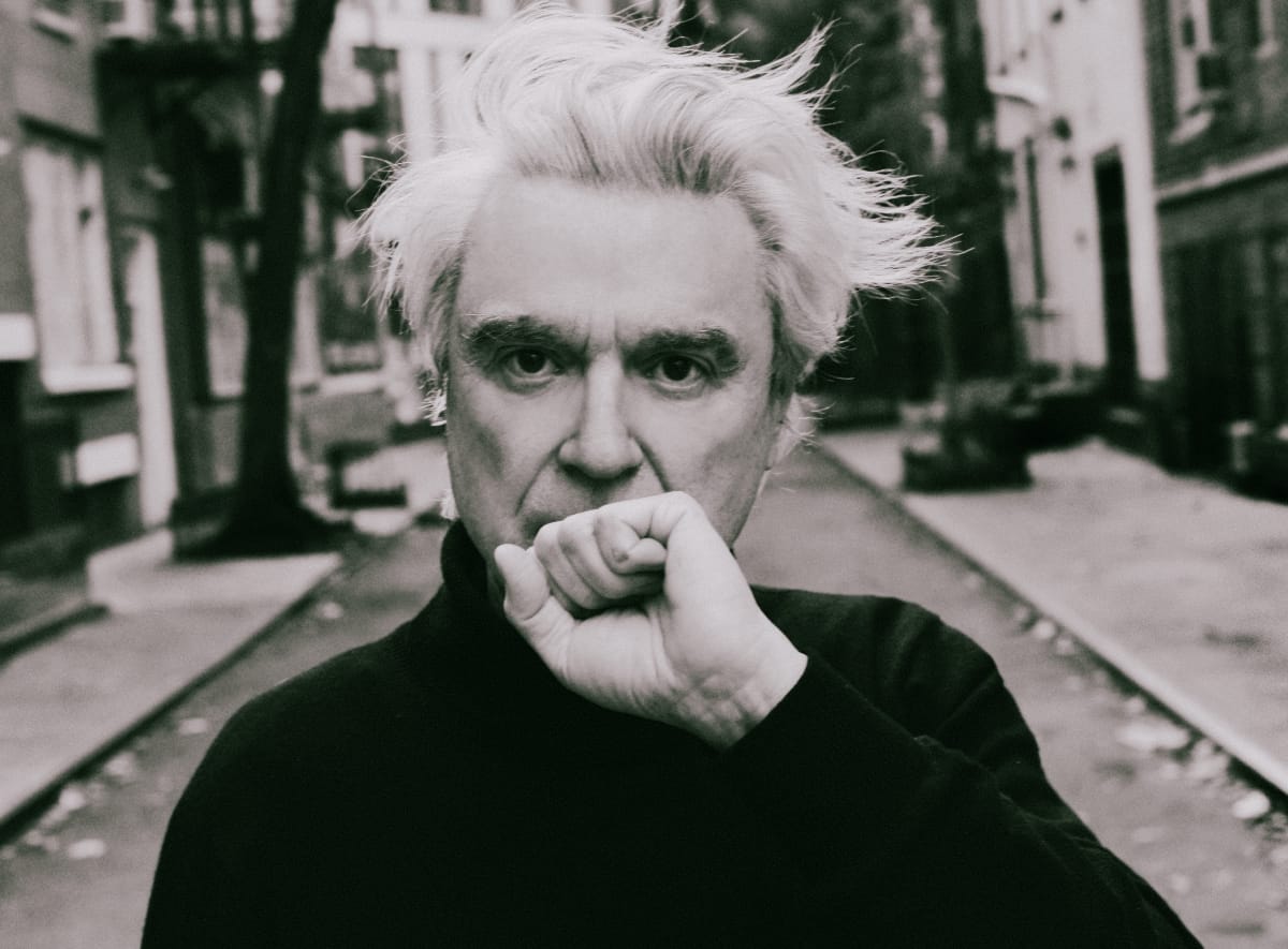 Black and white photo of David Byrne with his hand (or fist) in front of his mouth, looking serious. Photo by Shervin Lainez.