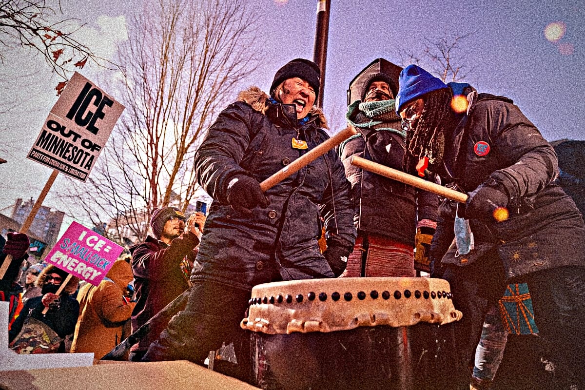 Protesters in winter gear play a large drum at Minneapolis anti-ICE demonstration, with "ICE OUT OF MINNESOTA" signs visible. Photograph by Lorie Shaull, courtesy of Wikimedia Commons.