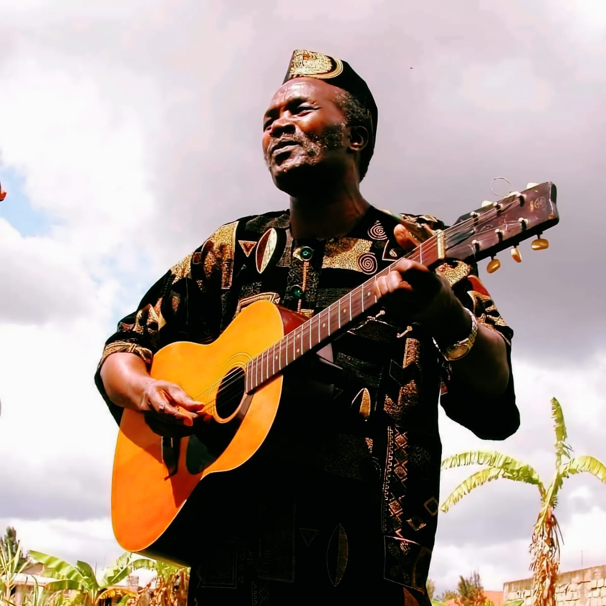 Joseph Kamaru plays acoustic guitar, photographed from below against a cloudy sky. He wears traditional patterned clothing and an embellished cap. Photo courtesy of Estate of Joseph Kamaru.