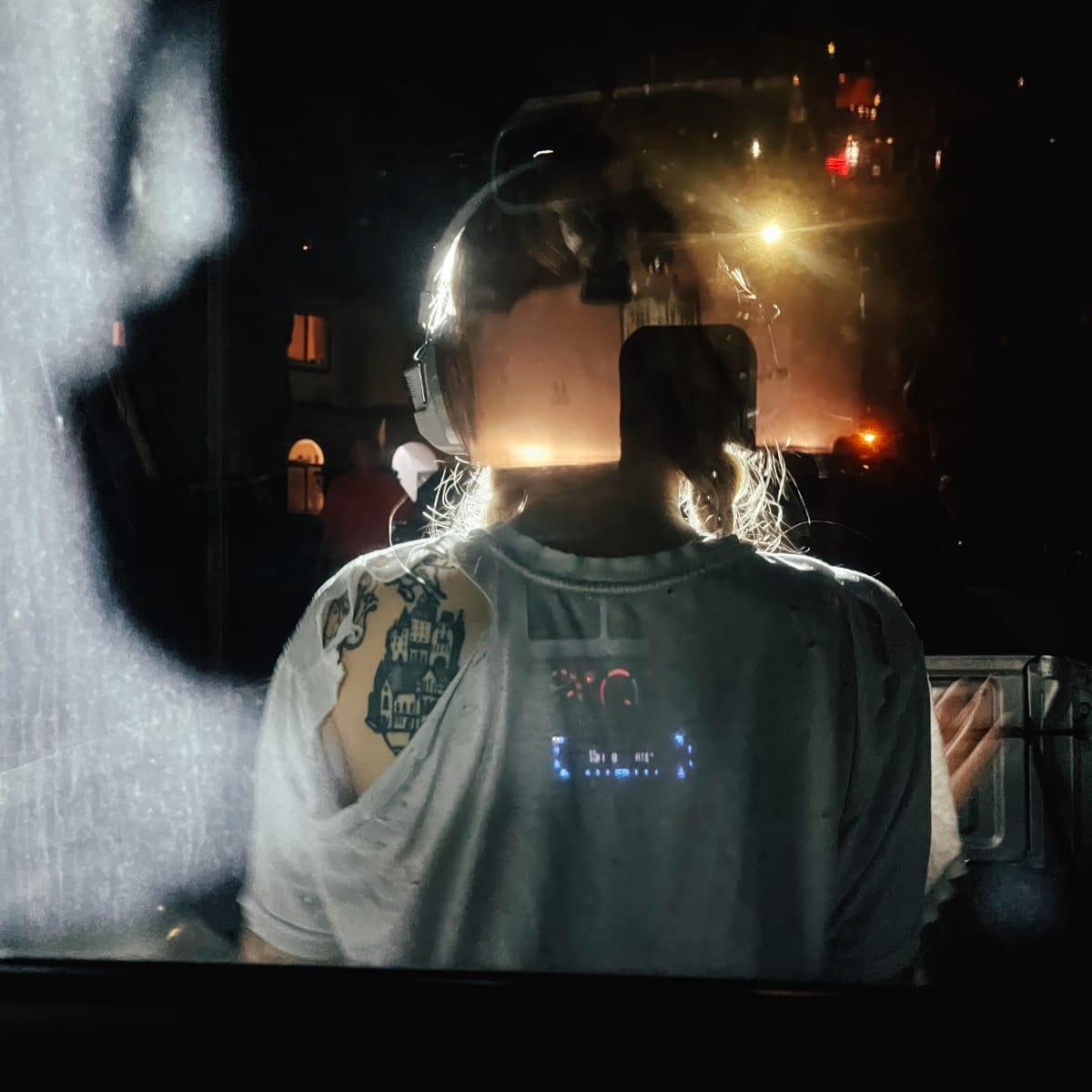 Lala Lala from behind wearing headphones and graphic t-shirt, facing illuminated screen in dark setting with ambient lights.