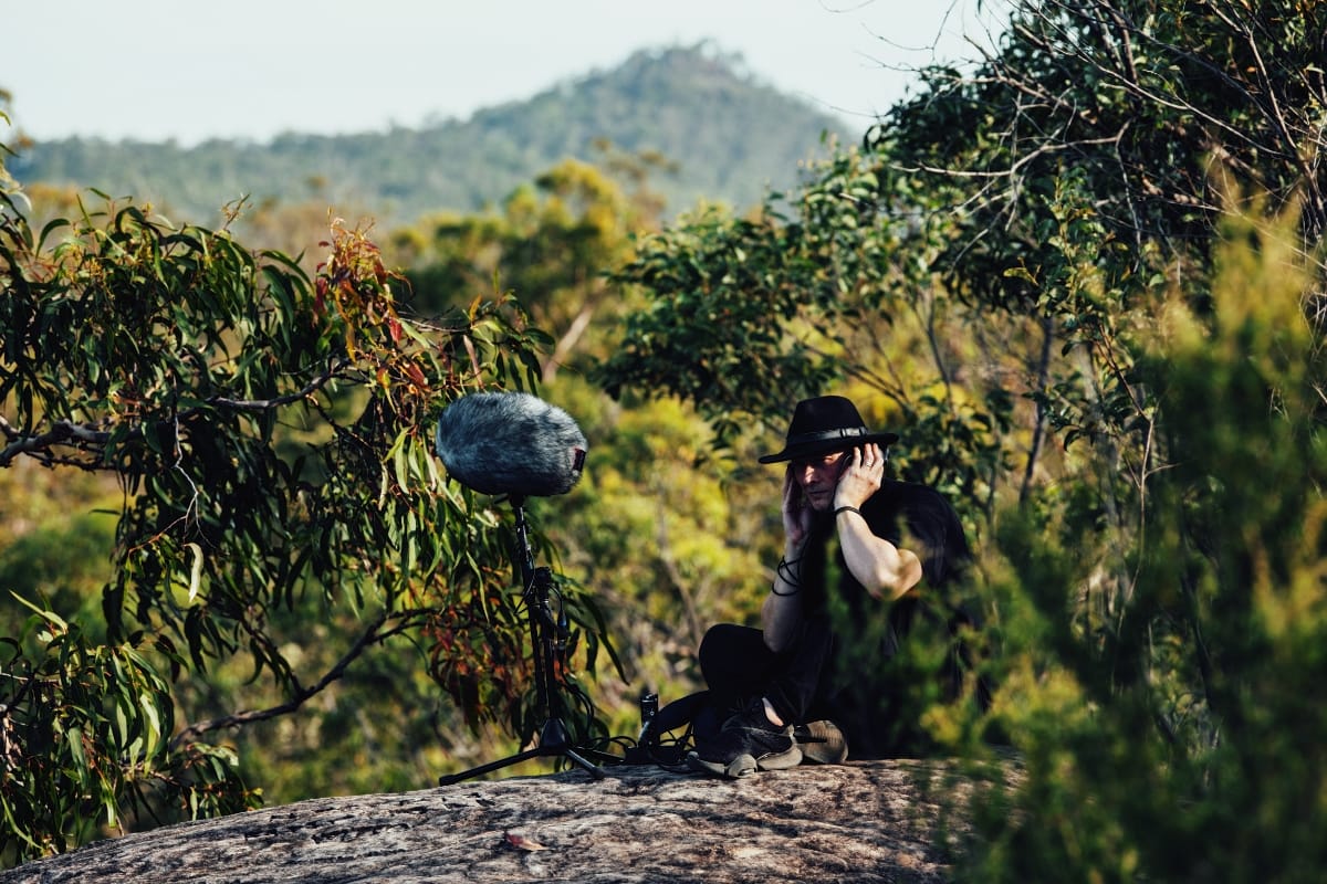 Lawrence English crouches on a rock outcrop with recording equipment, capturing field sounds in the lush Australian landscape of Nugum. Photo by Greg Harms.