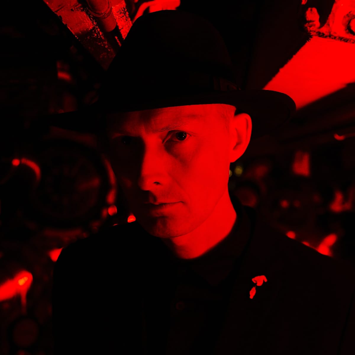 Lawrence English bathed in vivid red light against darkness, creating a stark, dramatic portrait with equipment visible in background. Photo by by D Wright.