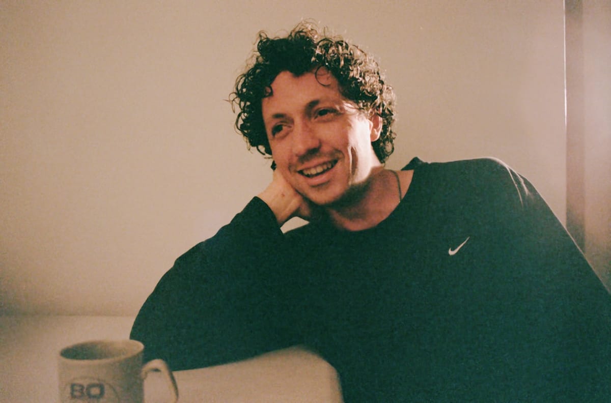 Liam Kazar smiles at the camera with his hand raised near his head. He has curly dark hair and wears a teal sweatshirt. A white mug sits nearby. Photo by Alexa Viscius.