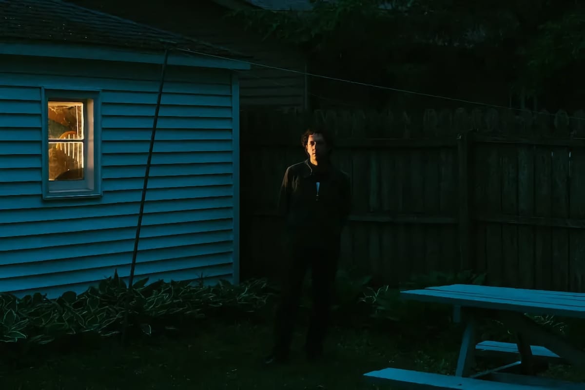 Liam Kazar, in dark clothing, stands by a teal structure with a lit window at twilight, surrounded by overgrown grass and foliage in blue-green light. Photo by Alexa Viscius