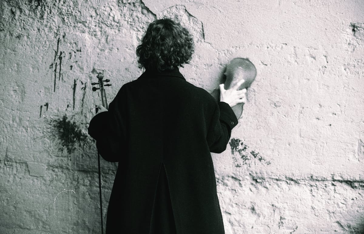 Marija Kovačević from behind with violin against textured white wall, black and white photograph showing curly dark hair. Photo by Anton Trofymov.