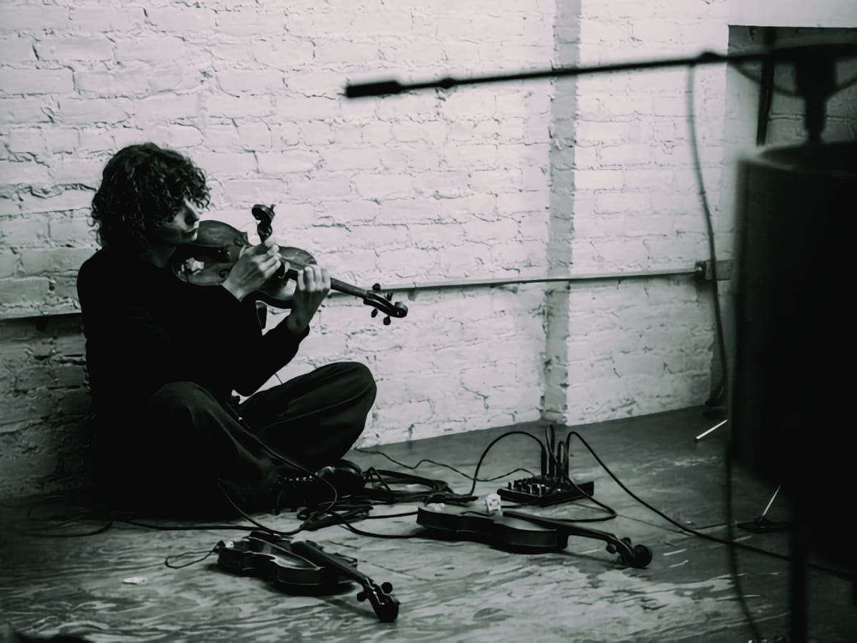 Marija Kovačević sits on floor playing violin surrounded by electronic equipment and cables against white brick wall, black and white. Photo by Peter Gannushkin.