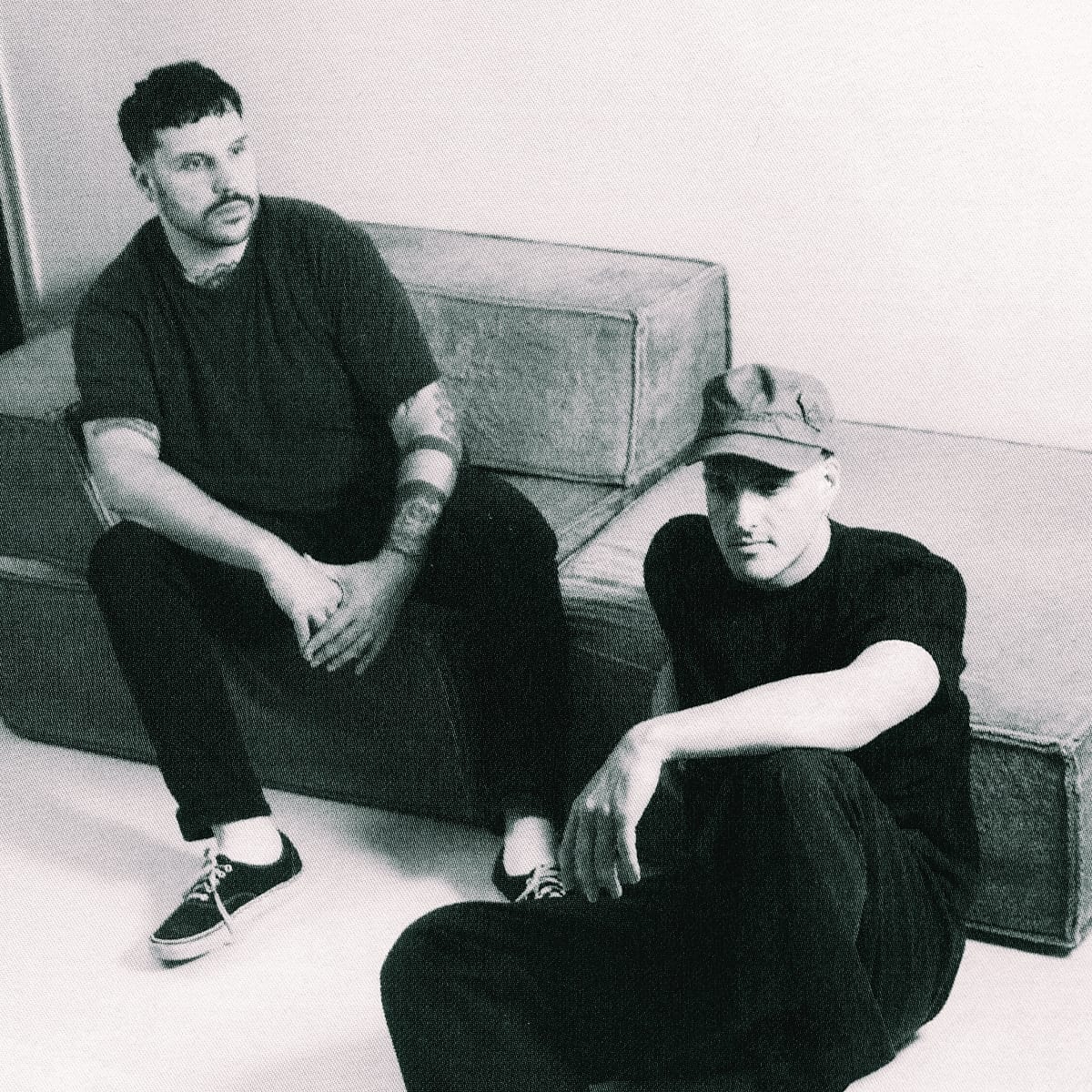 Max Scholl and Dhanya Langer of Modha in a minimal interior. Scholl sits on furniture, Langer on floor in cap, both wearing black.