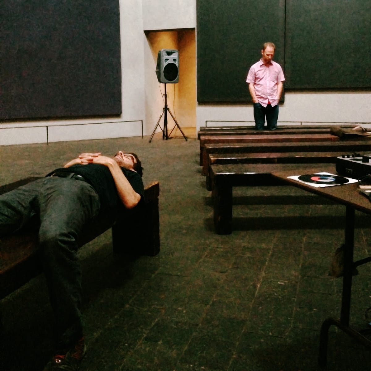 Stephen Vitiello lies on a bench during soundcheck at Rothko Chapel, Steve Roden stands in background, 2012. Large dark panels on walls. Photo by Sari Roden.