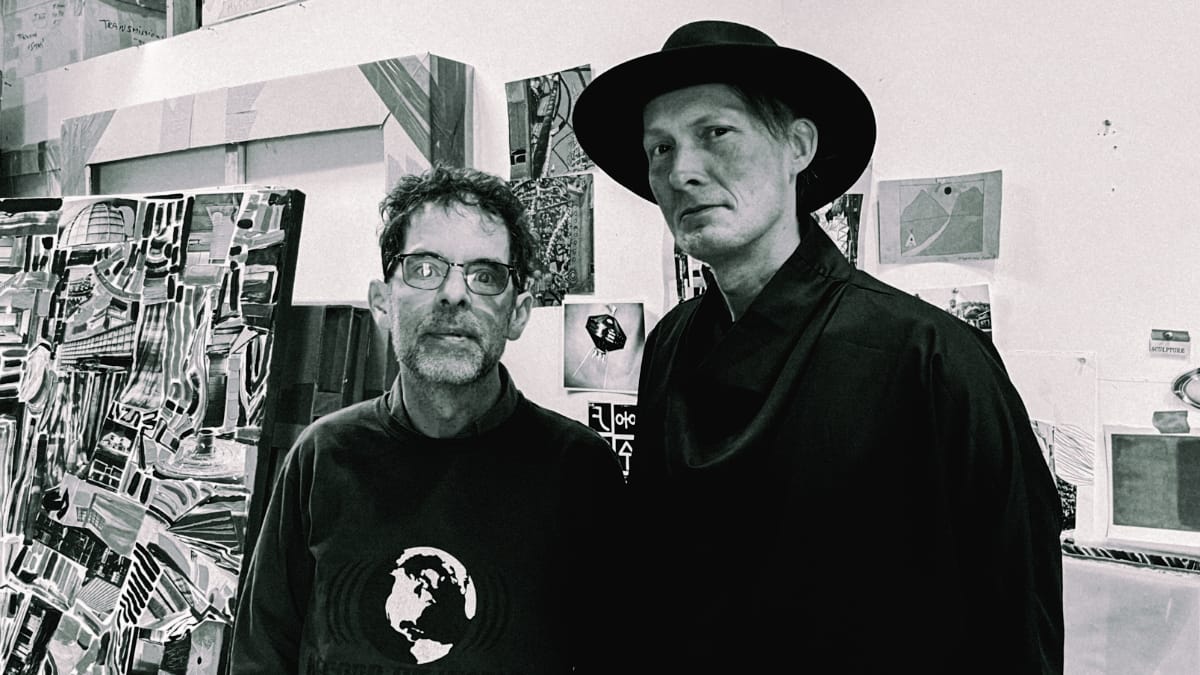Stephen Vitiello and Lawrence English in studio space, English wears black hat. Artwork and prints visible on walls, black and white.
