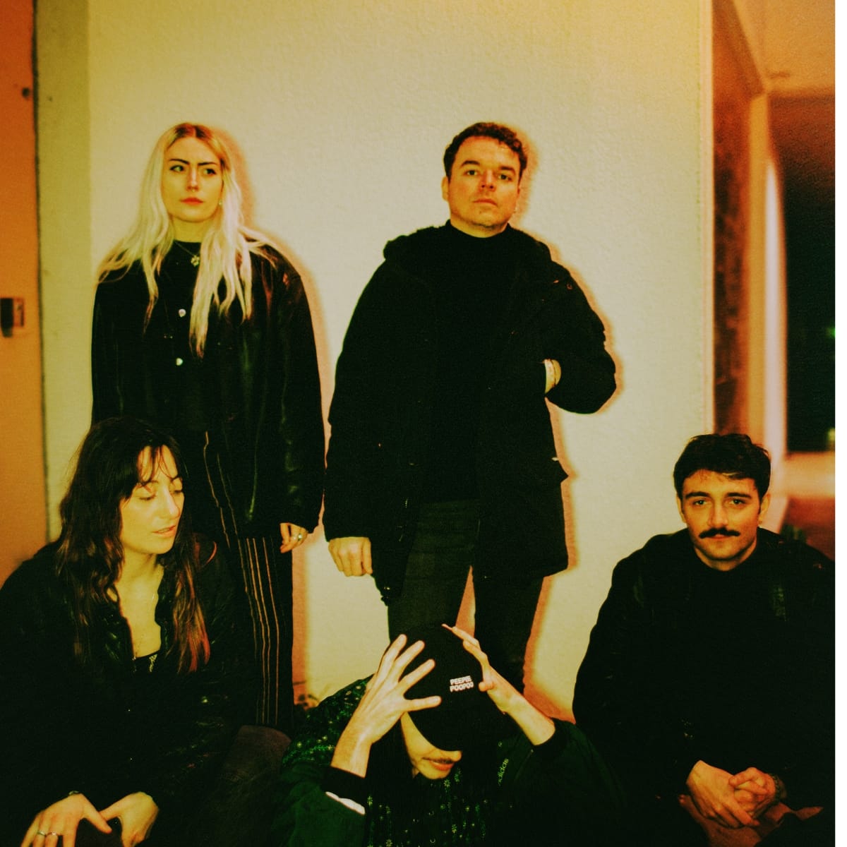 Four members of wavepool dressed in black pose against a white wall with warm lighting. Two stand while two sit in front. Photo by Charlotte Romer.