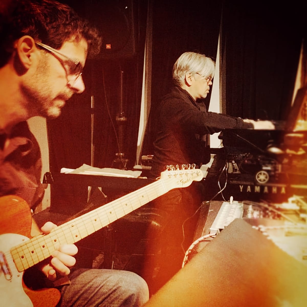 Stephen Vitiello on guitar in foreground, Ryuichi Sakamoto at Yamaha keyboard in background, 2012 performance with warm lighting. Photo by Taylor Deupree.