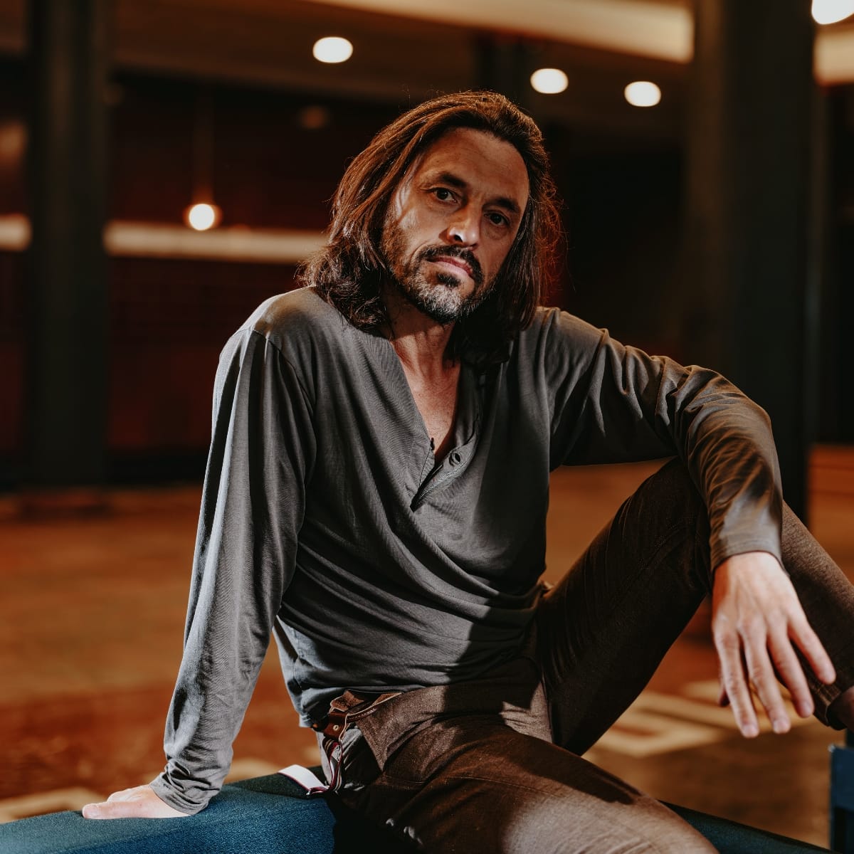 Aukai sits on wooden floor in studio space with warm lighting, shoulder-length dark hair, beard, wearing gray shirt. Photo by Jan Dellügge.