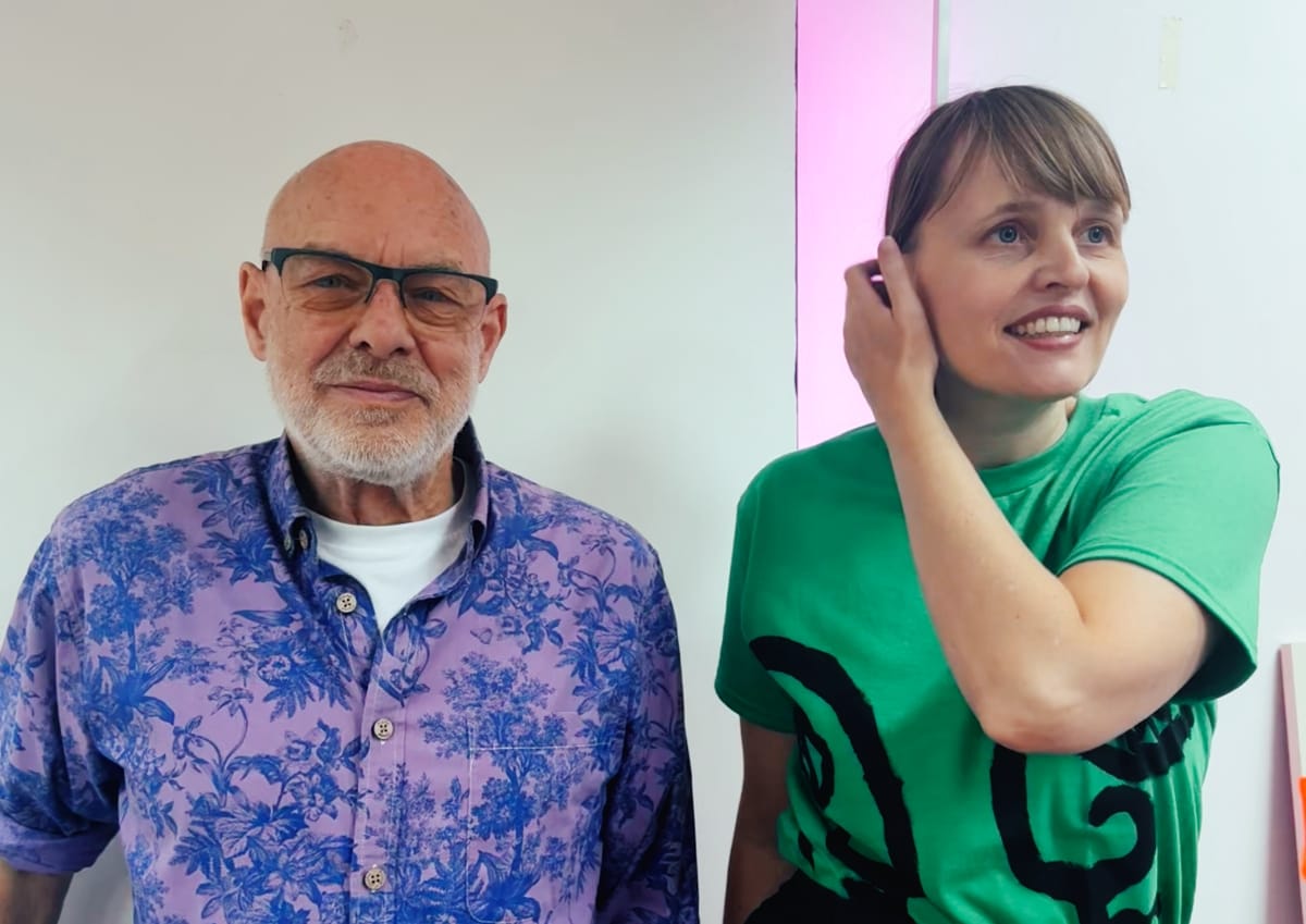 Brian Eno in blue floral shirt and Bette A in green patterned dress pose together, pink accent visible in background.