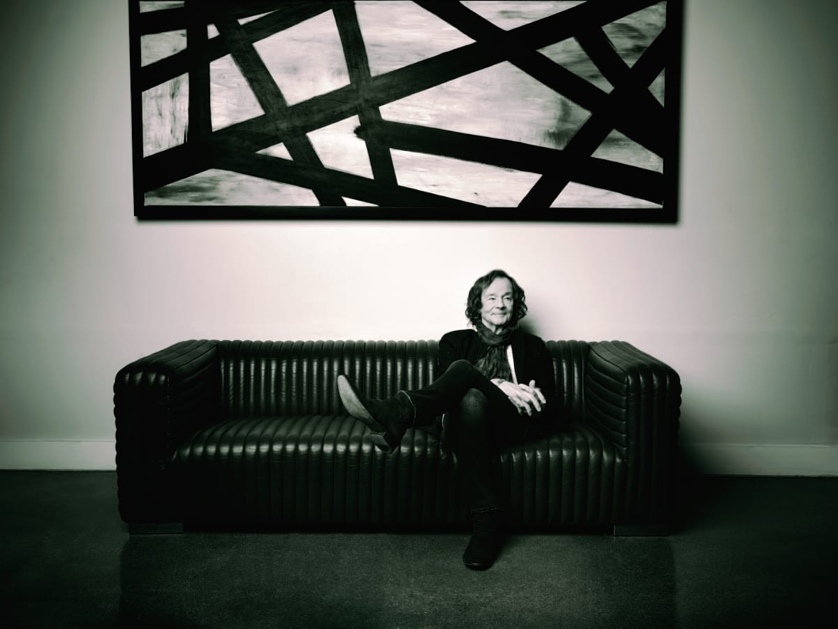 Colin Blunstone seated on ribbed black couch beneath geometric abstract artwork, wearing dark clothing and scarf, black and white tones.
