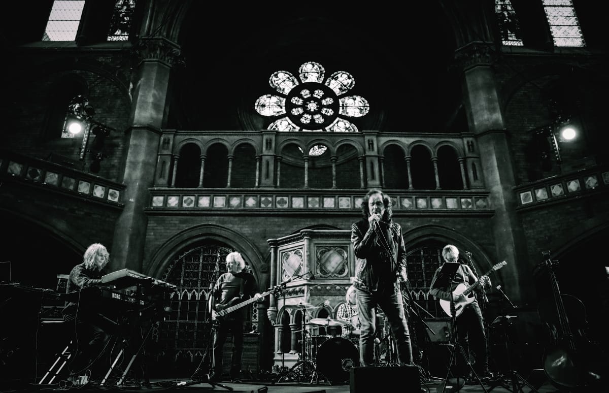 Colin Blunstone performs center stage at Union Chapel, band flanking, dramatic rose window and ornate architecture behind, black and white.