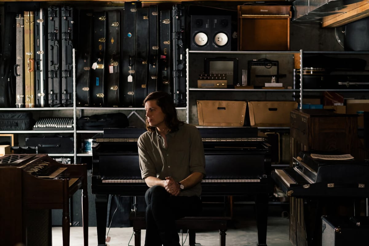 Erik Hall sits at piano in equipment-filled studio space, surrounded by shelves of instruments and gear, dramatic lighting. Photo by Carolina Mariana Rodriguez.