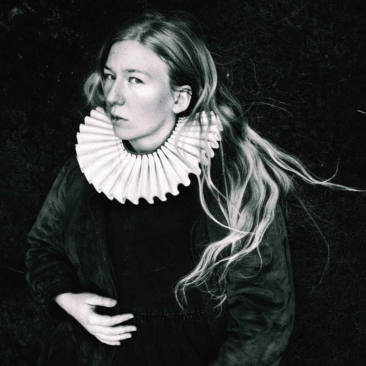 Fågelle in black and white portrait wearing white pleated ruff collar and dark clothing against dark natural background. Photo by Lars Bylund.