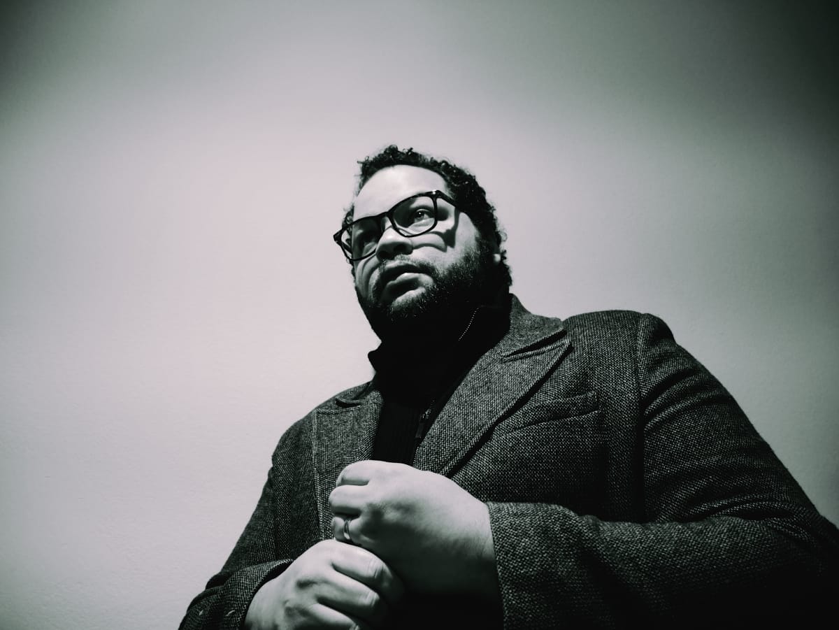 Juanma Trujillo in black and white low-angle portrait, wearing glasses and textured jacket against gradient background. Photo by Leonor Falcón.