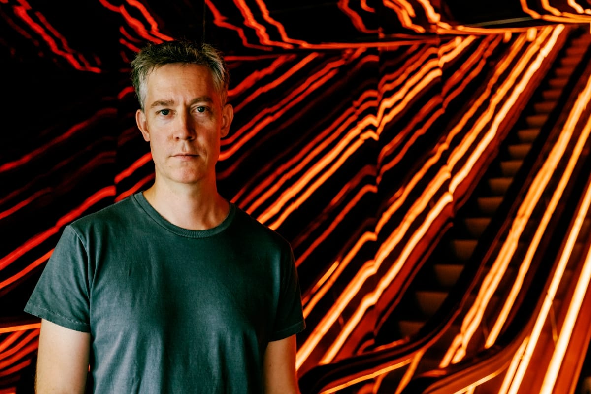 Kodomo in green t-shirt against dramatic flowing orange and red light patterns on black background.