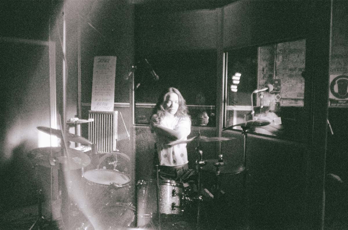 Momoko Gill at drums in grainy black and white studio photo, equipment and posters visible in background. Photo by Sophia Poole.