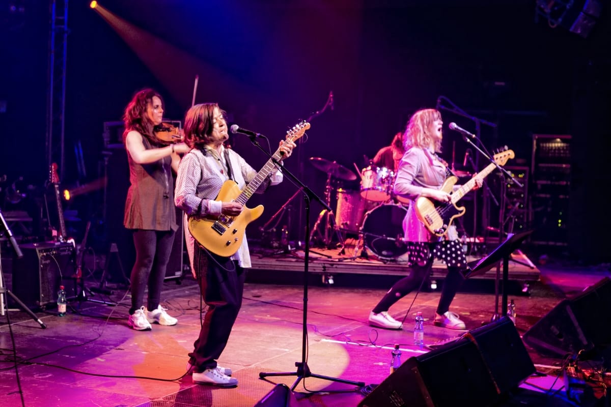 The Raincoats perform at Donau Festival, 2009, with violin, guitars, and drums under purple-pink stage lighting. Photo by Florian Schulte and courtesy The Raincoats.