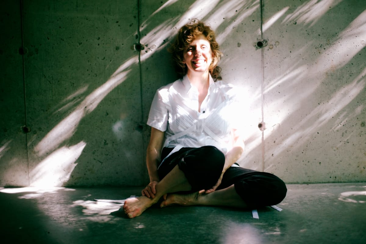 Zeena Parkins sits cross-legged on green floor in white shirt, curly hair, against textured wall with dramatic diagonal shadows. Photo by Jeff Preiss.