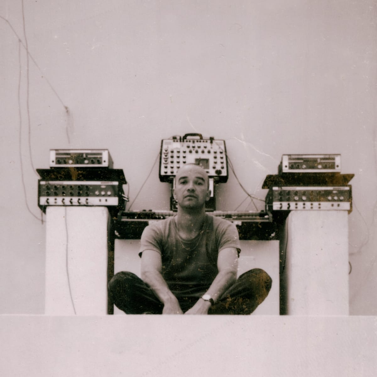 A vintage sepia photo of a shaved-head man (Conrad Schnitzler) seated cross-legged on the floor, surrounded by early electronic equipment and synthesizers arranged symmetrically on either side.