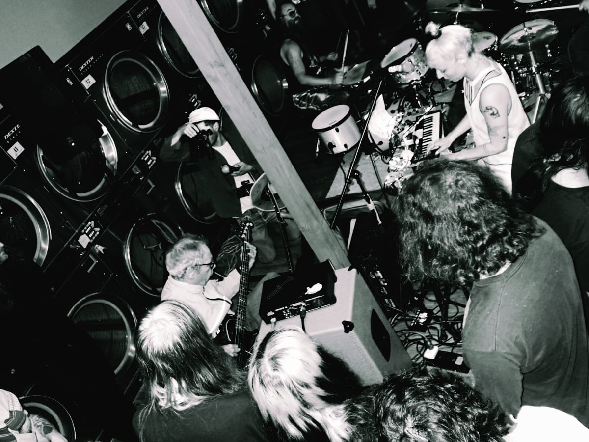 Galecstasy and Mike Watt Trio perform in a packed laundromat; bassist, vocalist, drummer, and keyboardist play before a close crowd, Dexter washers lining the walls. Photo by Suraj Patra.
