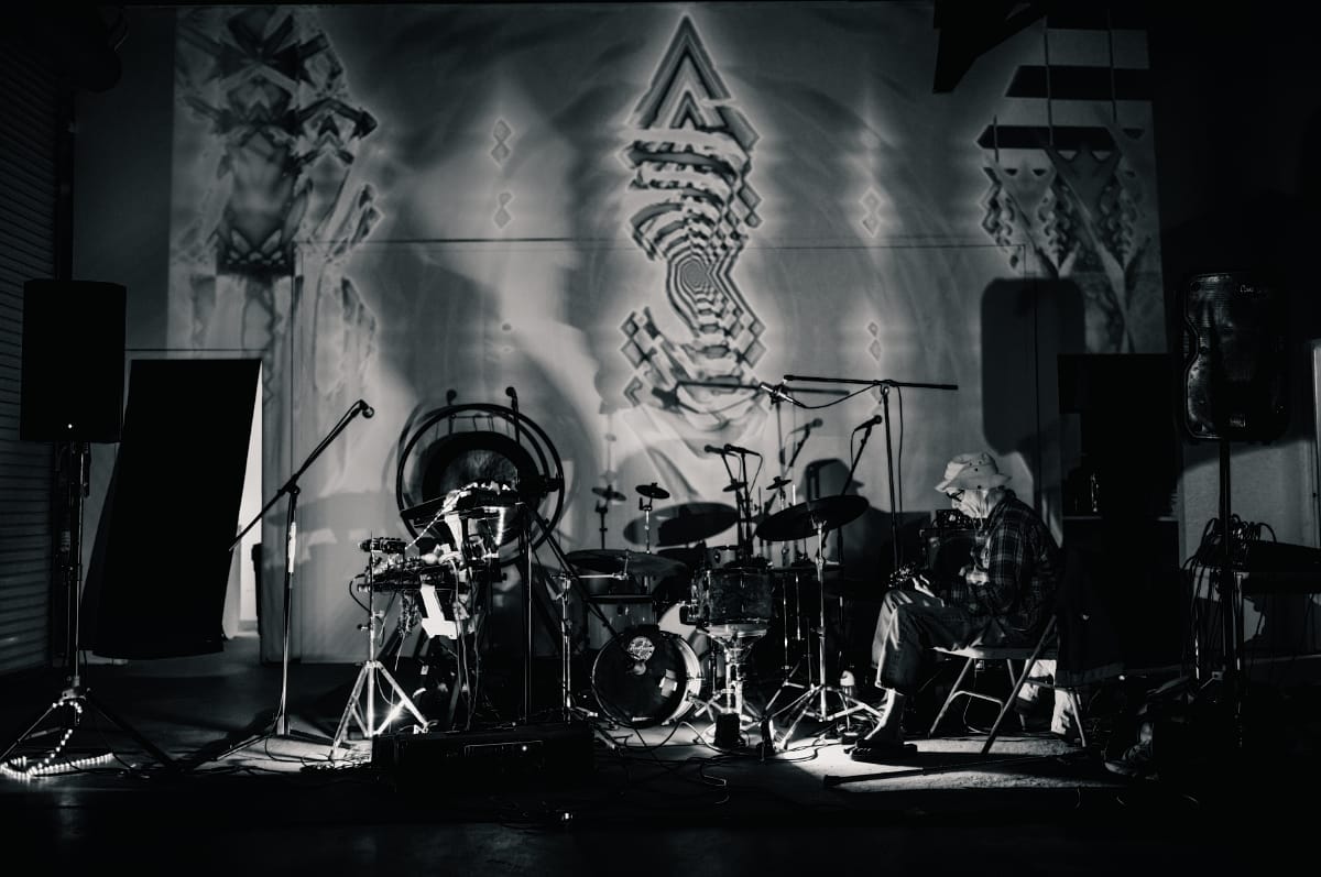 Mike Watt Trio performs on a darkened stage before a large psychedelic projected backdrop; a seated guitarist and drum kit are flanked by mic stands and PA speakers. Photo by Matthew Lawton.