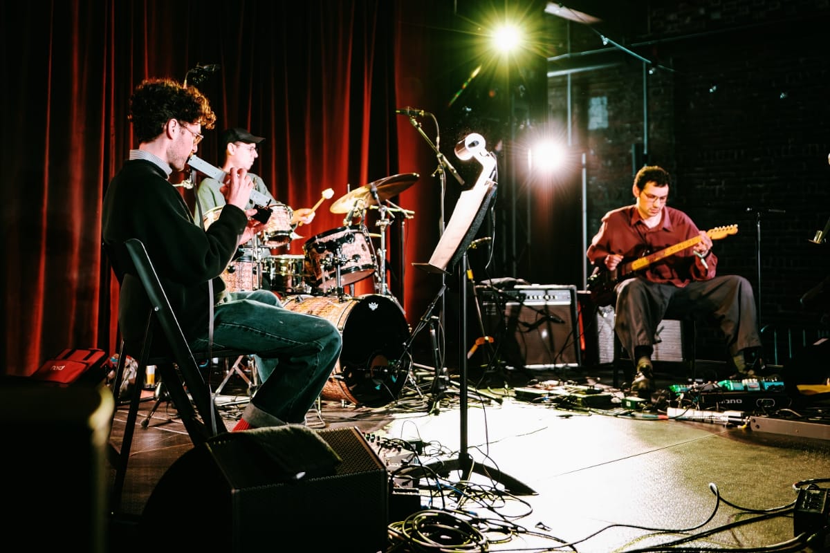 Three musicians (Gregory Uhlmann Trio) perform on a warmly lit stage with red curtains: a guitarist at left plays a wind instrument, a drummer plays center, and a second guitarist sits at right amid cables and pedals. Photo by Ash Ogle.
