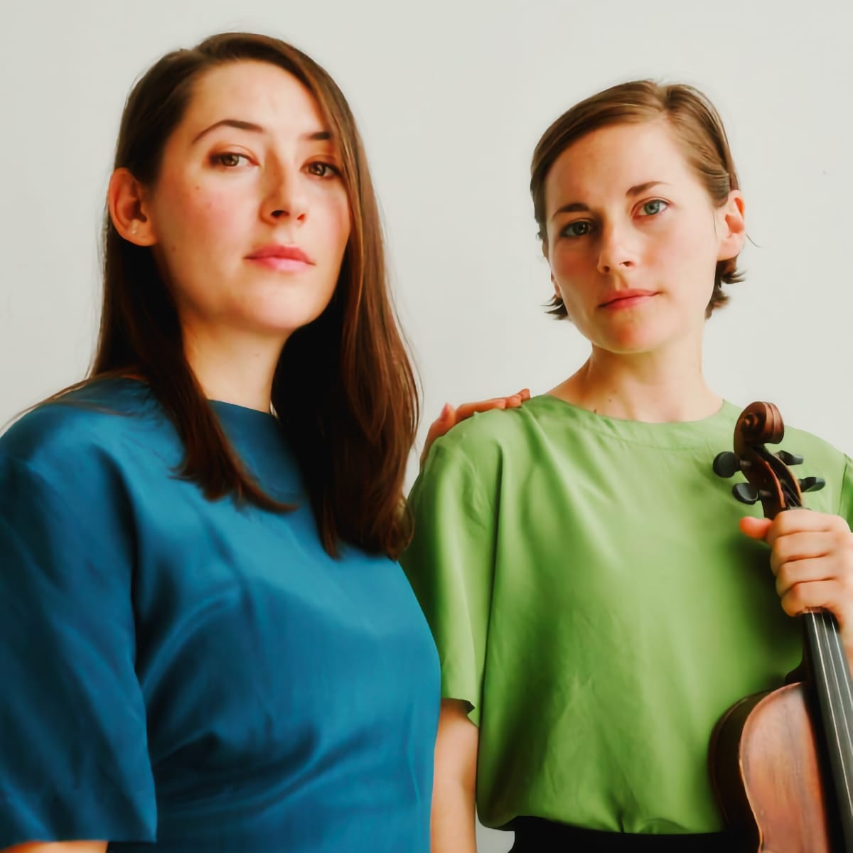 Two members of Sibyl side by side, one in blue shirt with long hair, one in green shirt holding violin, light background. Photo by Gregory Wikstrom.