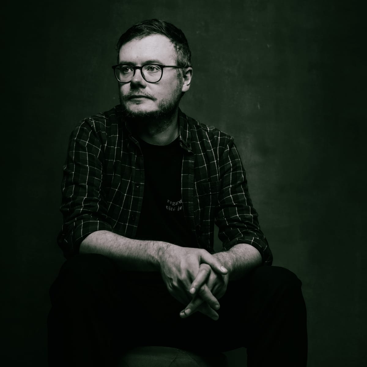 A moody studio portrait of Gavin Miller of worriedaboutsatan, seated with hands clasped, wearing round glasses and a plaid shirt against a dark green background. Photo by Jez Sheard.
