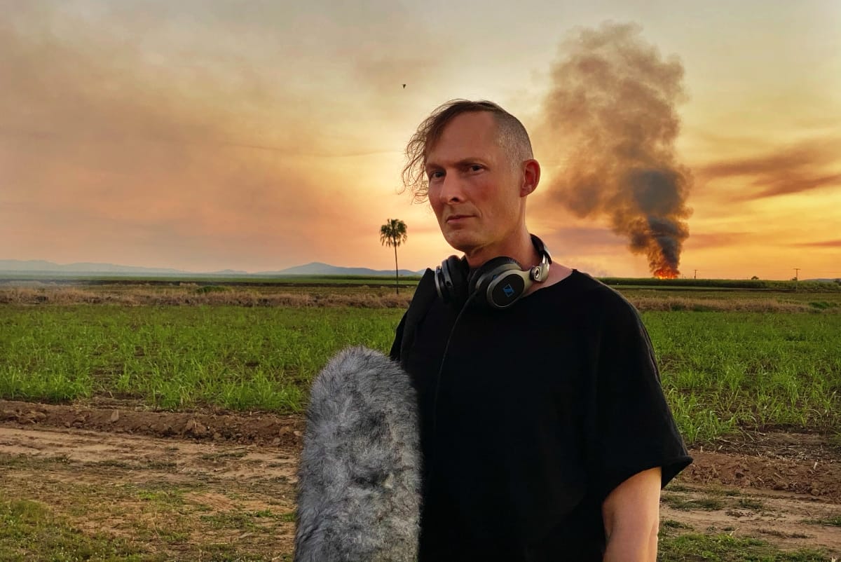 Lawrence English stands in an agricultural field with recording equipment and headphones, a plume of smoke rising in the distance at sunset. Photo by S Simpson.
