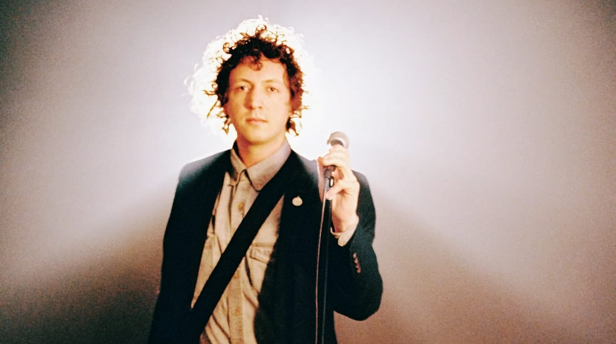 Liam Kazar holds a microphone, wearing a dark blazer. His backlit curly hair creates a halo effect. Soft gradient background, composed gaze. Photo by Alexa Viscius.