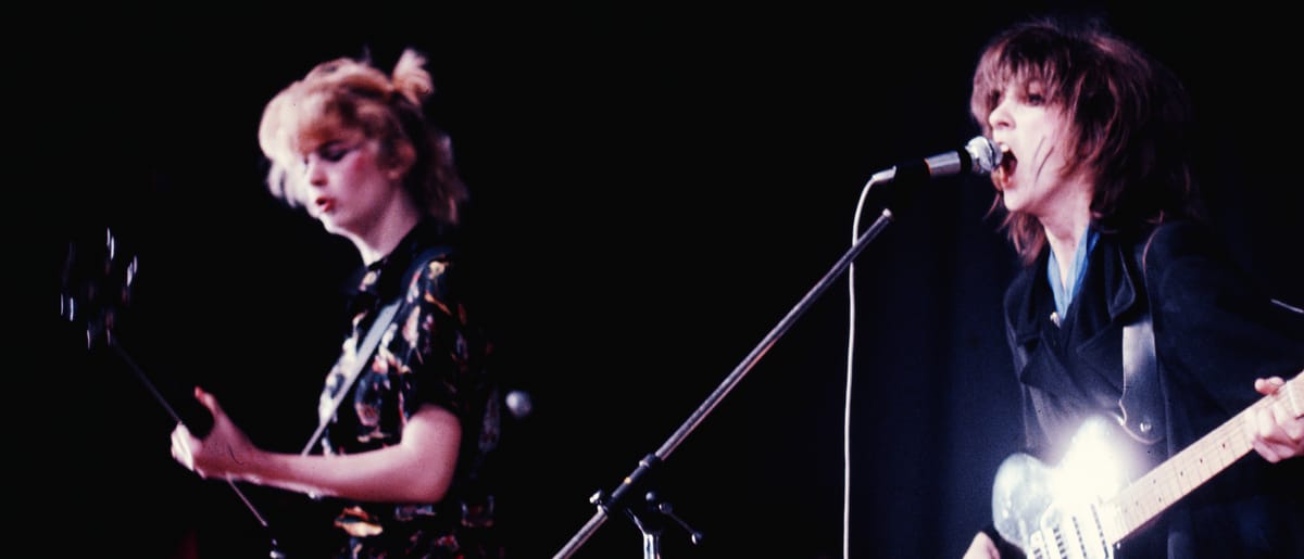 Gina and Ana of The Raincoats perform in Warsaw, 1978, vintage concert photo with guitar and microphone, purple-pink tones. Photo by Maria Helena da Silva and courtesy Ana da Silva.