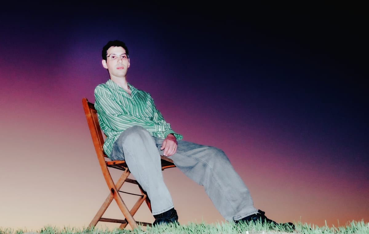 A young man (Gregory Uhlmann) in a green striped shirt and light jeans sits in a wooden folding chair on grass against a twilight gradient sky. Photo by Joyce Kim.