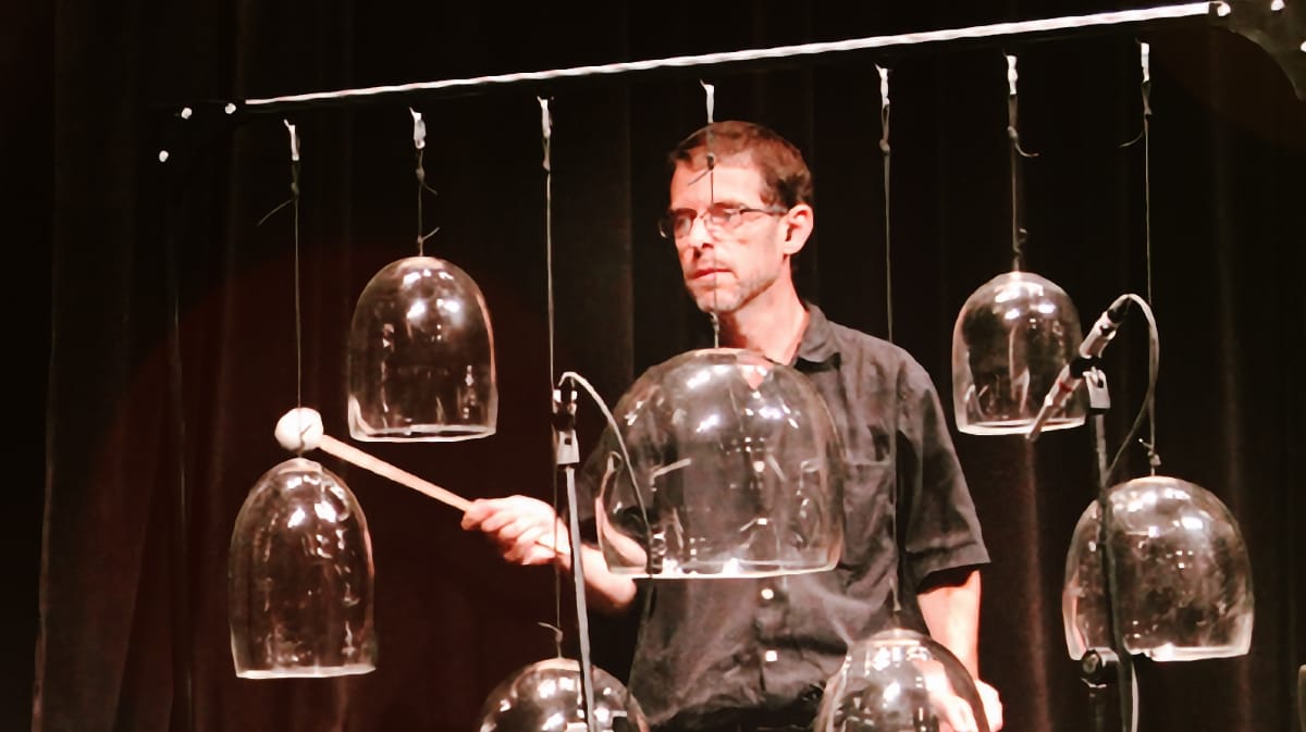 Stephen Vitiello at performance rehearsal with suspended glass bell instruments, holding a mallet against dark backdrop. Photo by Kevin Gafford, courtesy of the Museum of Modern Art, 2013.