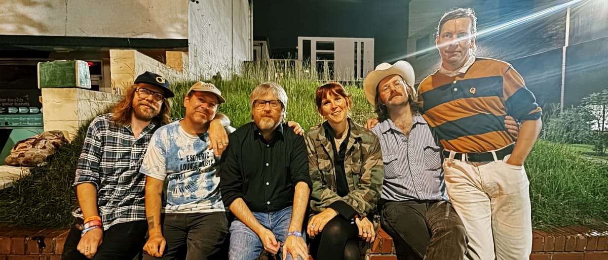 Six members of Winged Wheel pose together in casual attire, including hats and varied clothing, against a decorative prairie backdrop. Photo by Katy McElroy.
