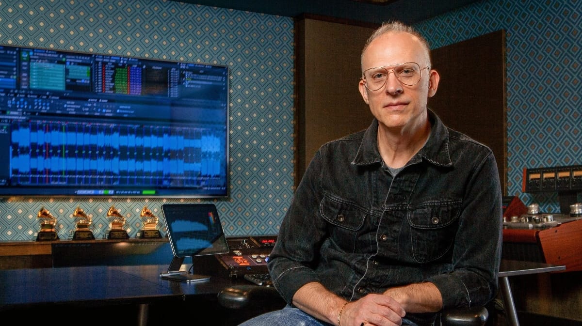 Michael Graves in recording studio wearing glasses and denim shirt, Grammy awards visible on desk, audio editing software on screen.
