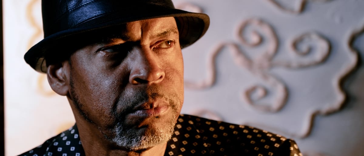 Bayeté (Todd Cochran) in hat and patterned shirt with warm dramatic lighting, decorative scrollwork visible in background.