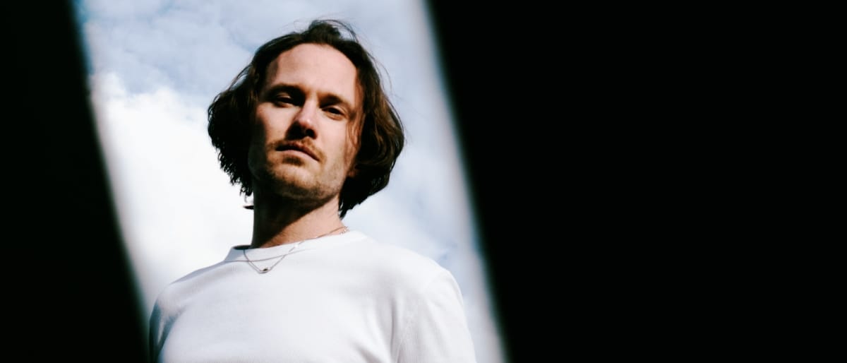 A man with shoulder-length hair (David August) and a white sweater is photographed from below against a cloudy sky, framed by dark vertical shapes. Photo by Sarah Fuchs.
