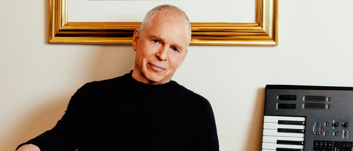 An older man (Peter Baumann) in a black turtleneck sits beside a keyboard synthesizer beneath a gold-framed artwork on a cream wall, looking directly at the camera. Photo by Jane Richey.
