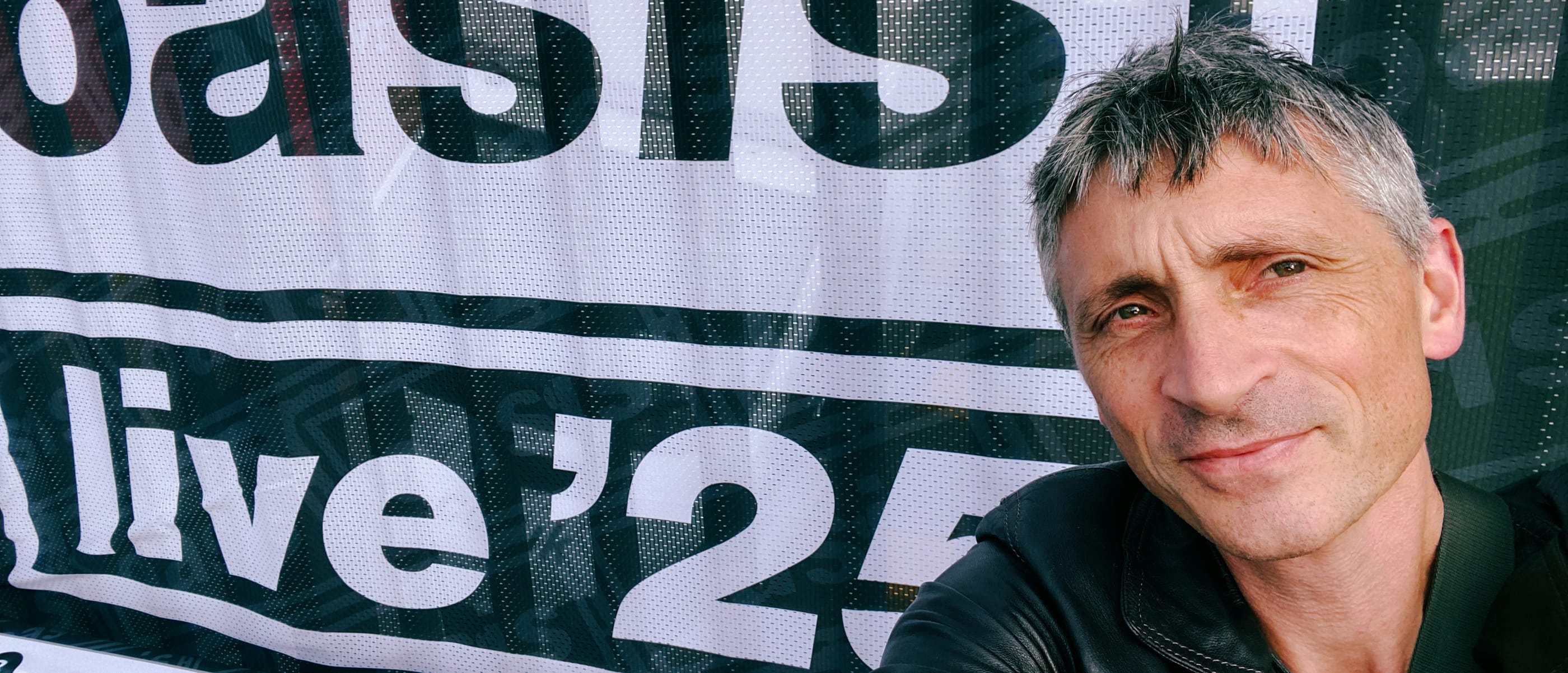 Ludo Hunter-Tilney poses in front of an Oasis "live '25" banner, wearing dark clothing with short gray hair.
