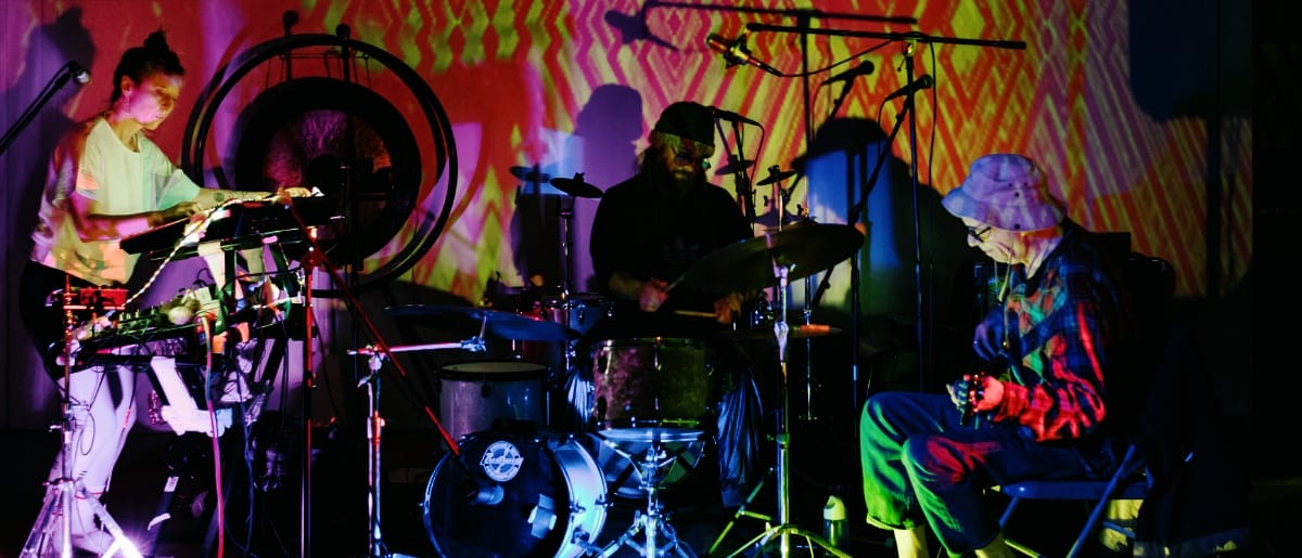 Galecstasy & Mike Watt Trio perform with psychedelic yellow-green projections, three musicians visible with electronic equipment and drums. Photo by Matthew Lawton.