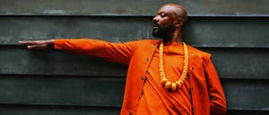 Justin Hicks in bright orange jacket with wooden bead necklace, arm extended against dark slatted wall background. Photo by Malik Well.