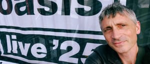 Ludo Hunter-Tilney poses in front of an Oasis "live '25" banner, wearing dark clothing with short gray hair.