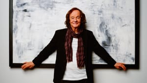 Colin Blunstone in black jacket and burgundy scarf against large abstract blue-white painting, arms outstretched touching frame.