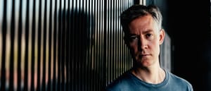 Kodomo in blue shirt against striped backdrop with moody lighting and subtle color accents.