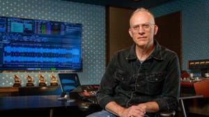 Michael Graves in recording studio wearing glasses and denim shirt, Grammy awards visible on desk, audio editing software on screen.