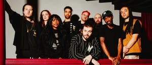 Nine members of Nubiyan Twist in dark clothing against white wall with dramatic shadows, arranged in group portrait with red foreground element.